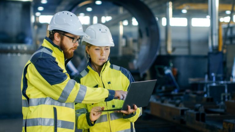 Dos trabajadores con casco revisando datos en un portátil en planta